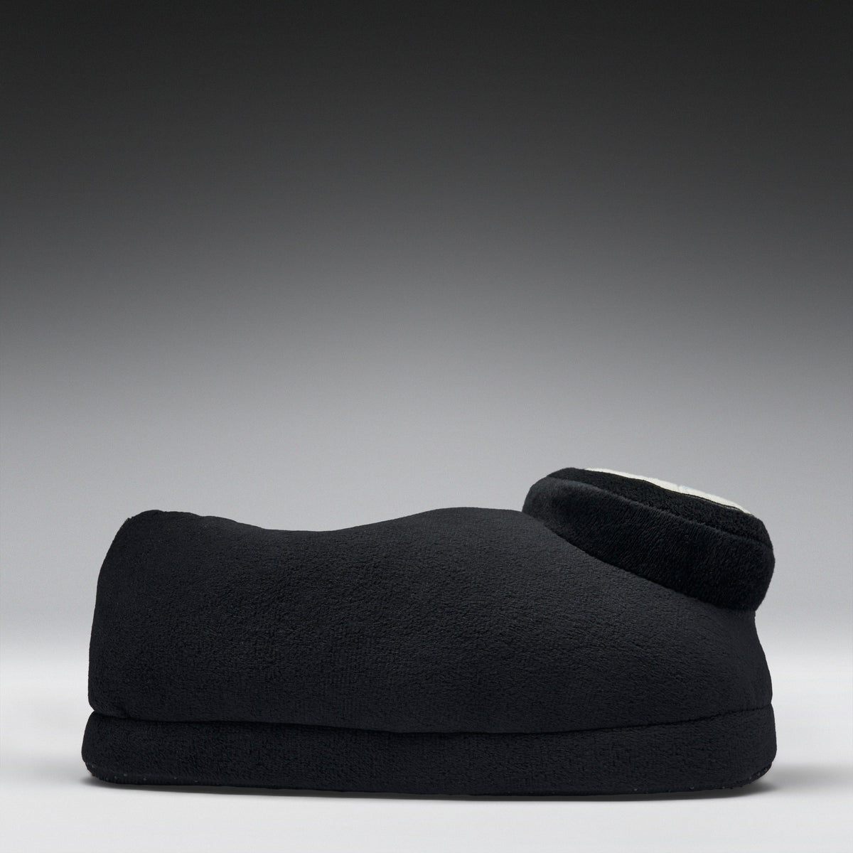 A black fleece-covered footrest or cushion with a curved shape and a small circular white accent on top, photographed against a dark gray to light gray gradient studio background.