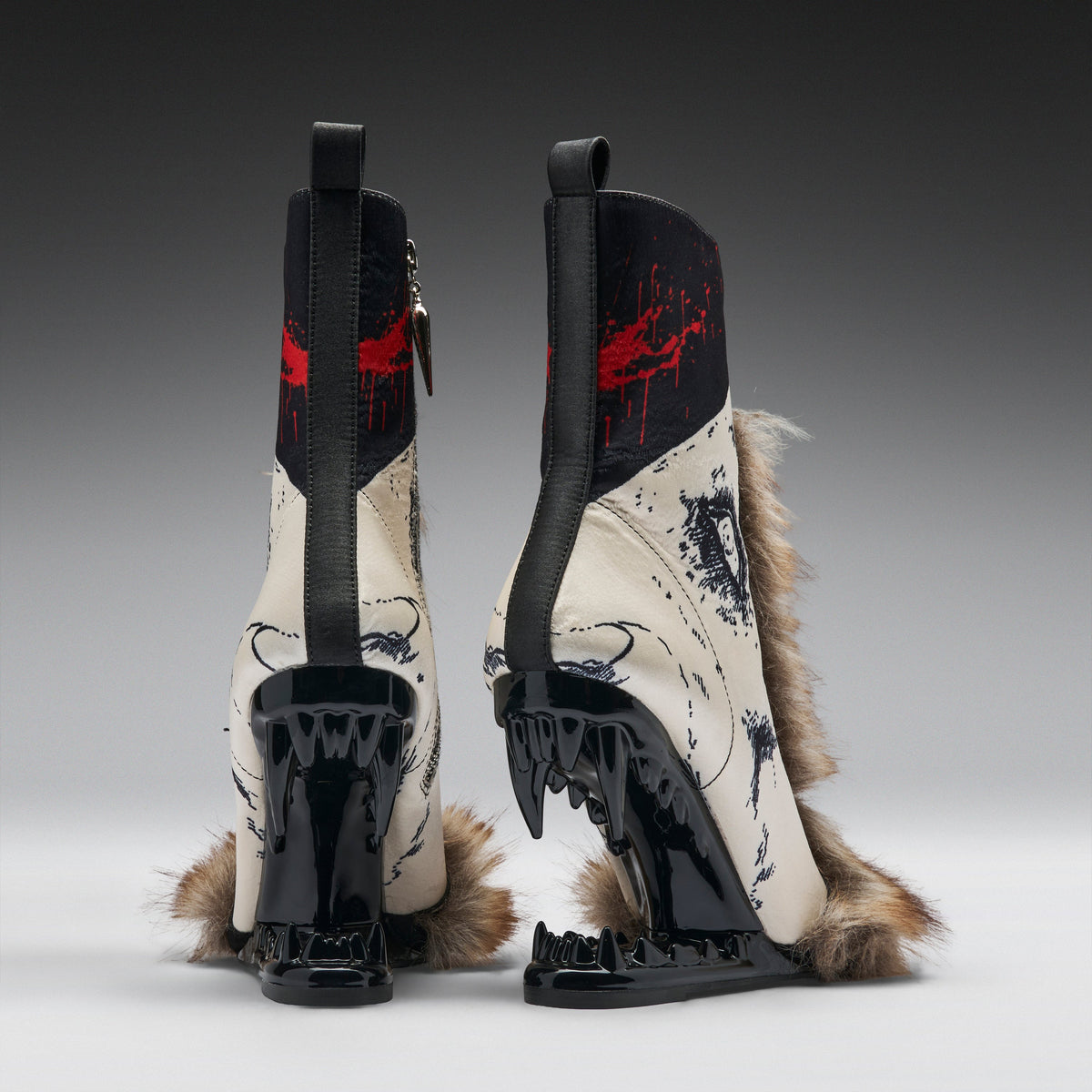 A pair of high-heeled boots shown from the back, featuring glossy black wedge heels with sharp teeth-like details, cream-colored panels with black graphic print, red paint splatter accents, and brown fur trim, photographed on a gray gradient studio background.