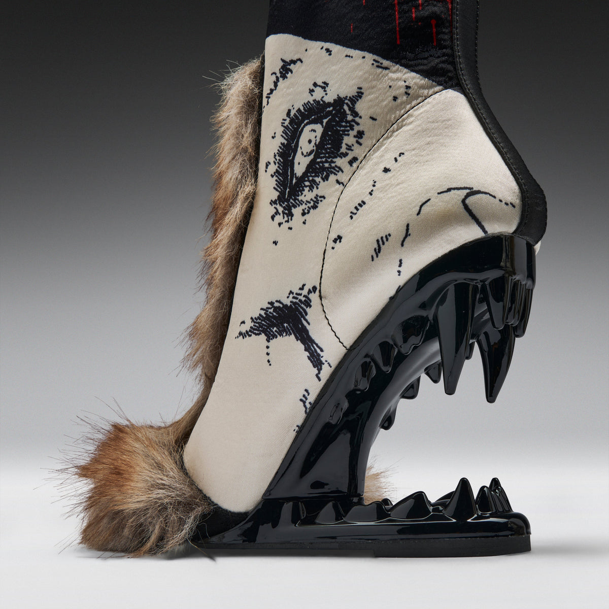 A single high-heeled boot shown in side profile on a studio background. The boot has a cream-colored upper with black graphic artwork, brown fur trim, and a glossy black sculptural heel and sole with sharp spike details.