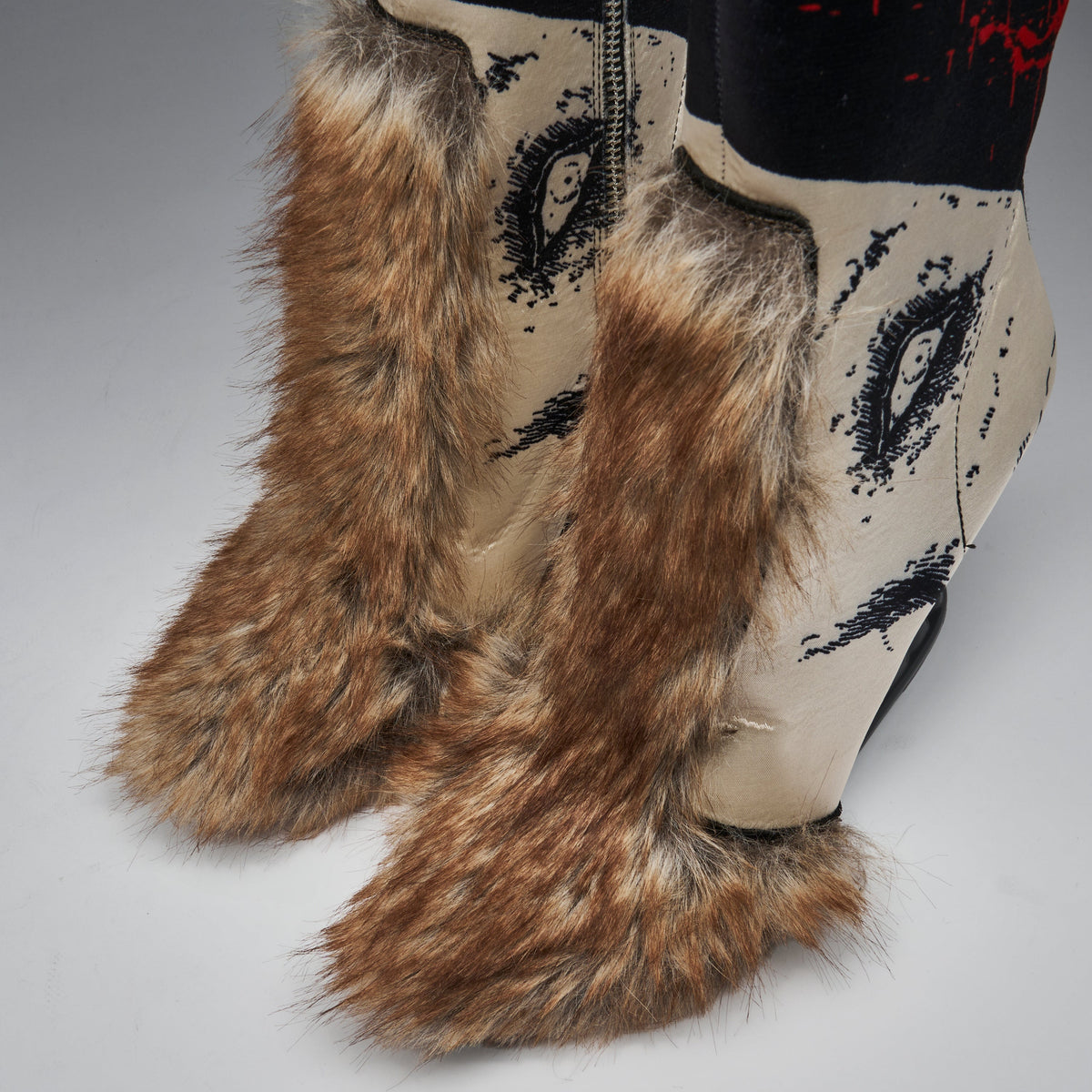 Close-up of a pair of western-style boots with long brown faux-fur leg warmers and foot covers, cream shafts with black eye-like graphics, and a black top section with red splatter details.