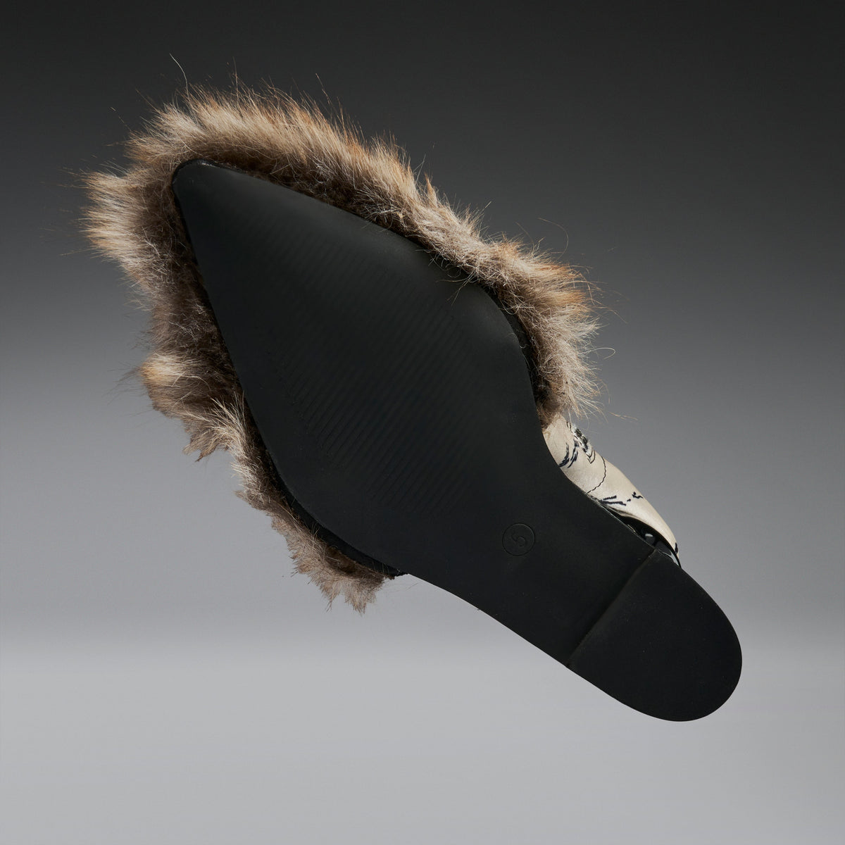A black slip-on shoe with a brown and beige faux-fur trim, shown from a side profile against a dark gray gradient background.