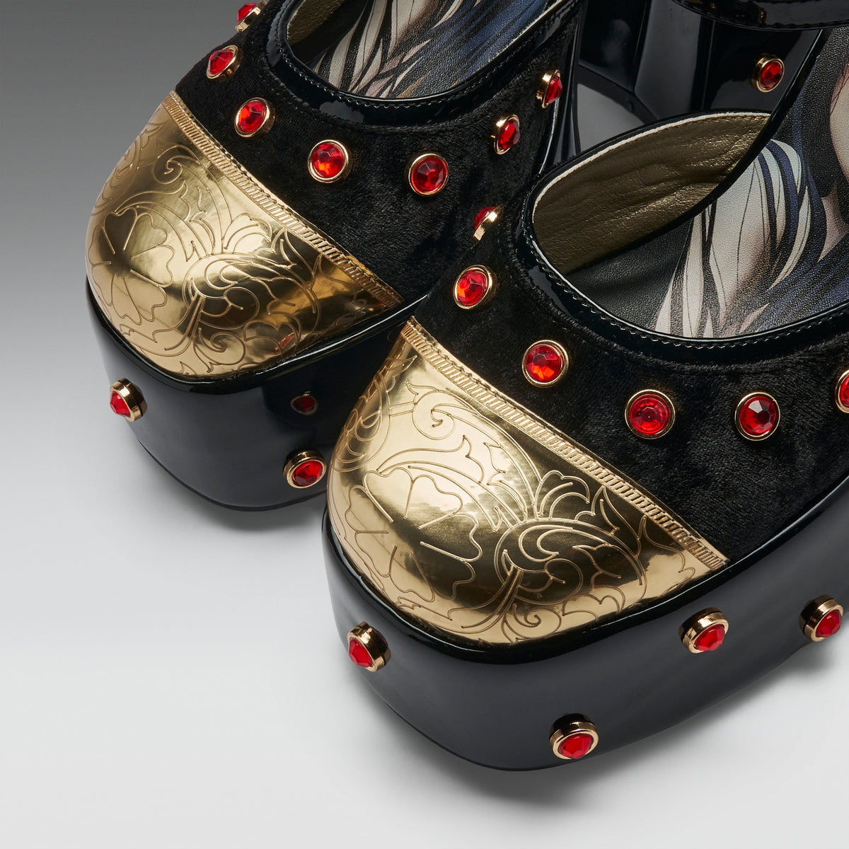 A close-up of a pair of platform Mary Jane shoes featuring black velvet uppers, metallic gold floral-embossed toe caps, black platform soles, and red rhinestone embellishments.
