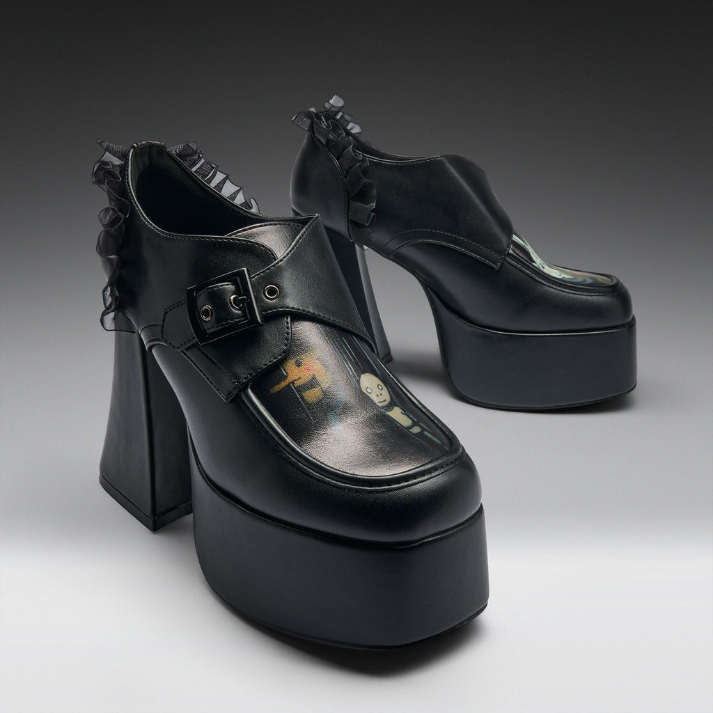 A pair of black platform mule heels with ruffled trim, shown on a clean gray studio background. The shoes feature a chunky block heel, a thick platform sole, a buckle strap across the top, and a glossy finish with a small printed graphic on the toe area.