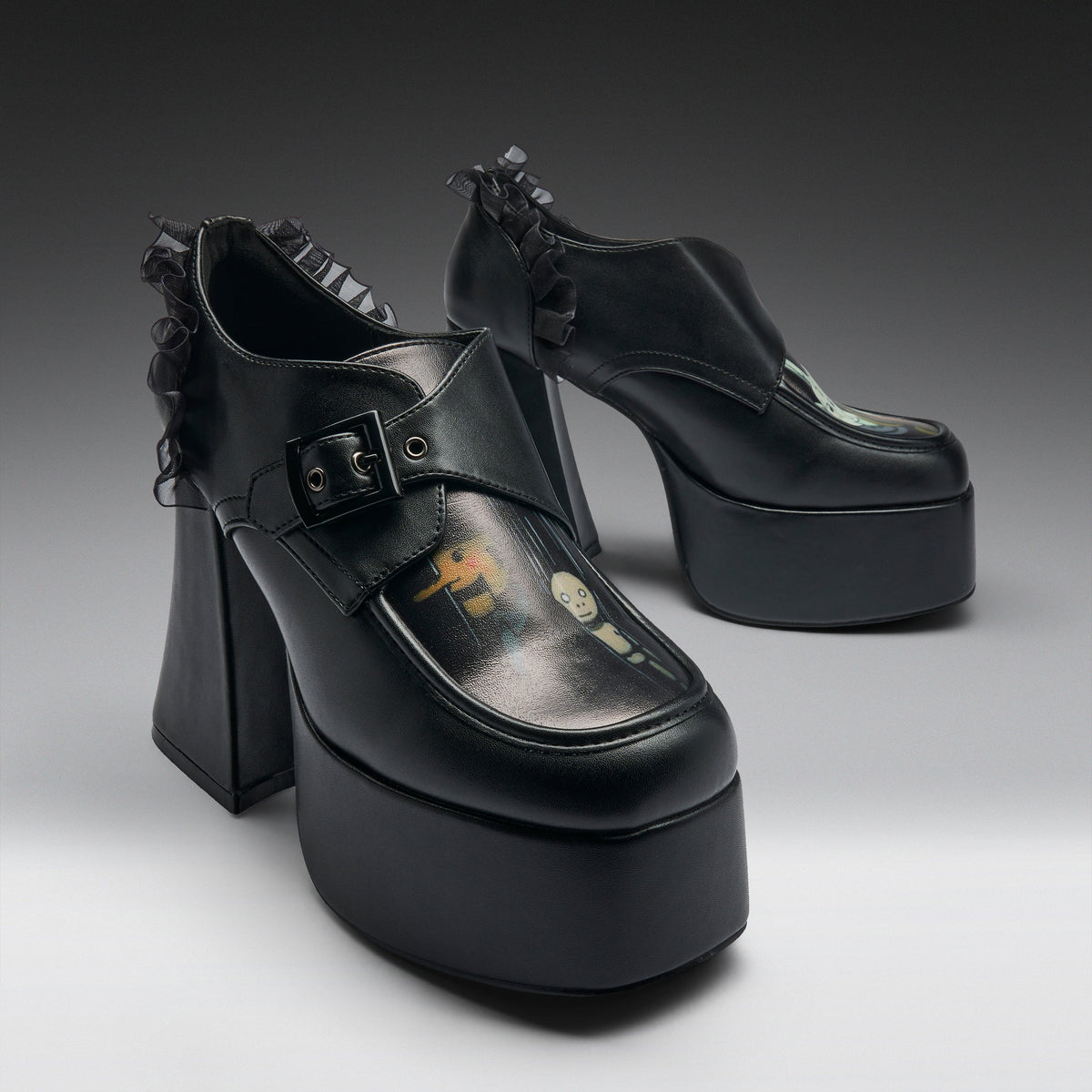 A pair of black platform mule heels with ruffled trim, shown on a clean gray studio background. The shoes feature a chunky block heel, a thick platform sole, a buckle strap across the top, and a glossy finish with a small printed graphic on the toe area.