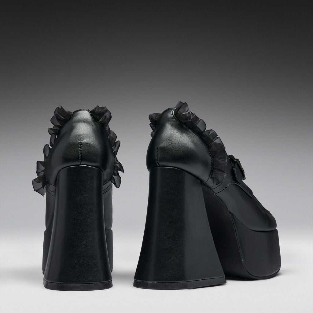 A pair of black platform high heels with dramatic ruffled detailing on the sides, shown from the back against a dark gray gradient studio background.