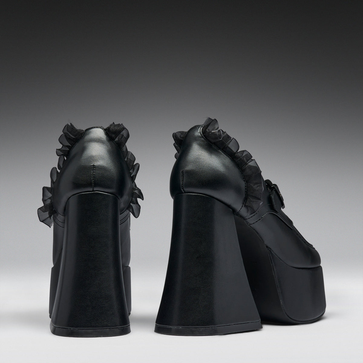 A pair of black platform high heels with dramatic ruffled detailing on the sides, shown from the back against a dark gray gradient studio background.