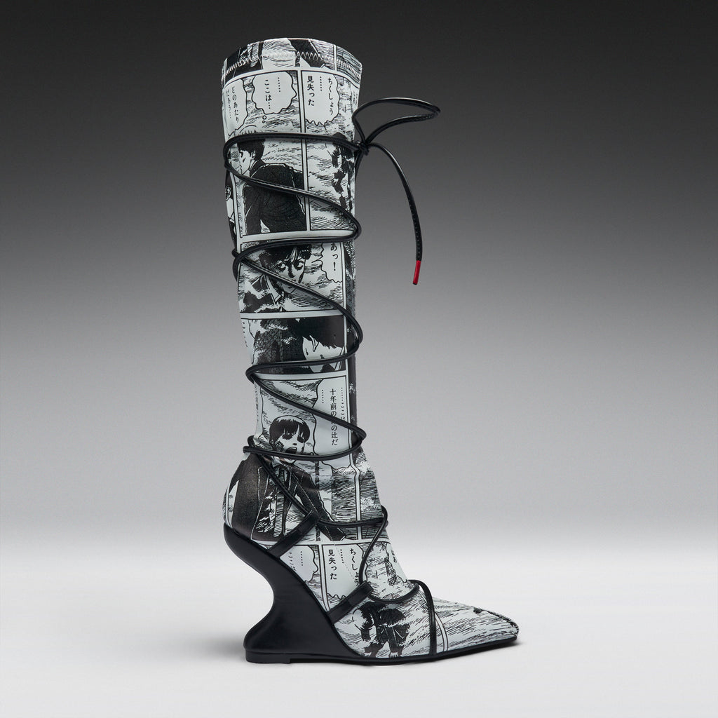 A single tall knee-high boot with a black and white manga comic book print, wrapped in black lace up to the ankle, set against a dark-to-light gray gradient studio background.