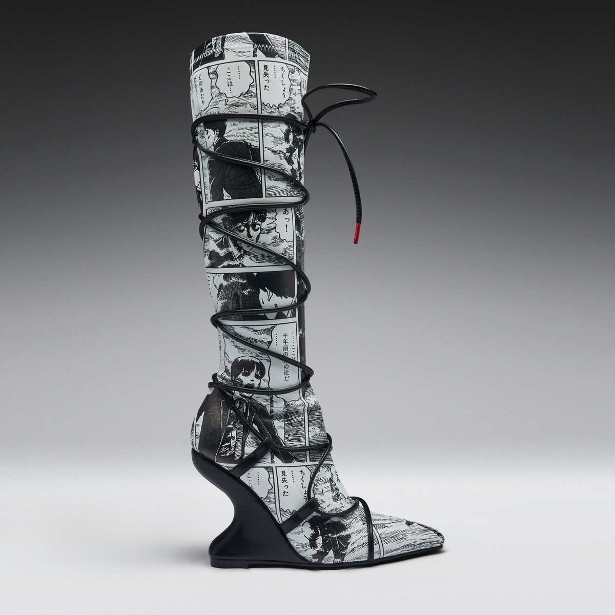 A single tall knee-high boot with a black and white manga comic book print, wrapped in black lace up to the ankle, set against a dark-to-light gray gradient studio background.