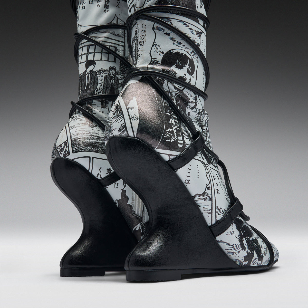 A pair of high-heeled boots with sculptural black wedge heels and black straps, featuring a black-and-white manga comic book print on the uppers, shown against a dark gray studio background.
