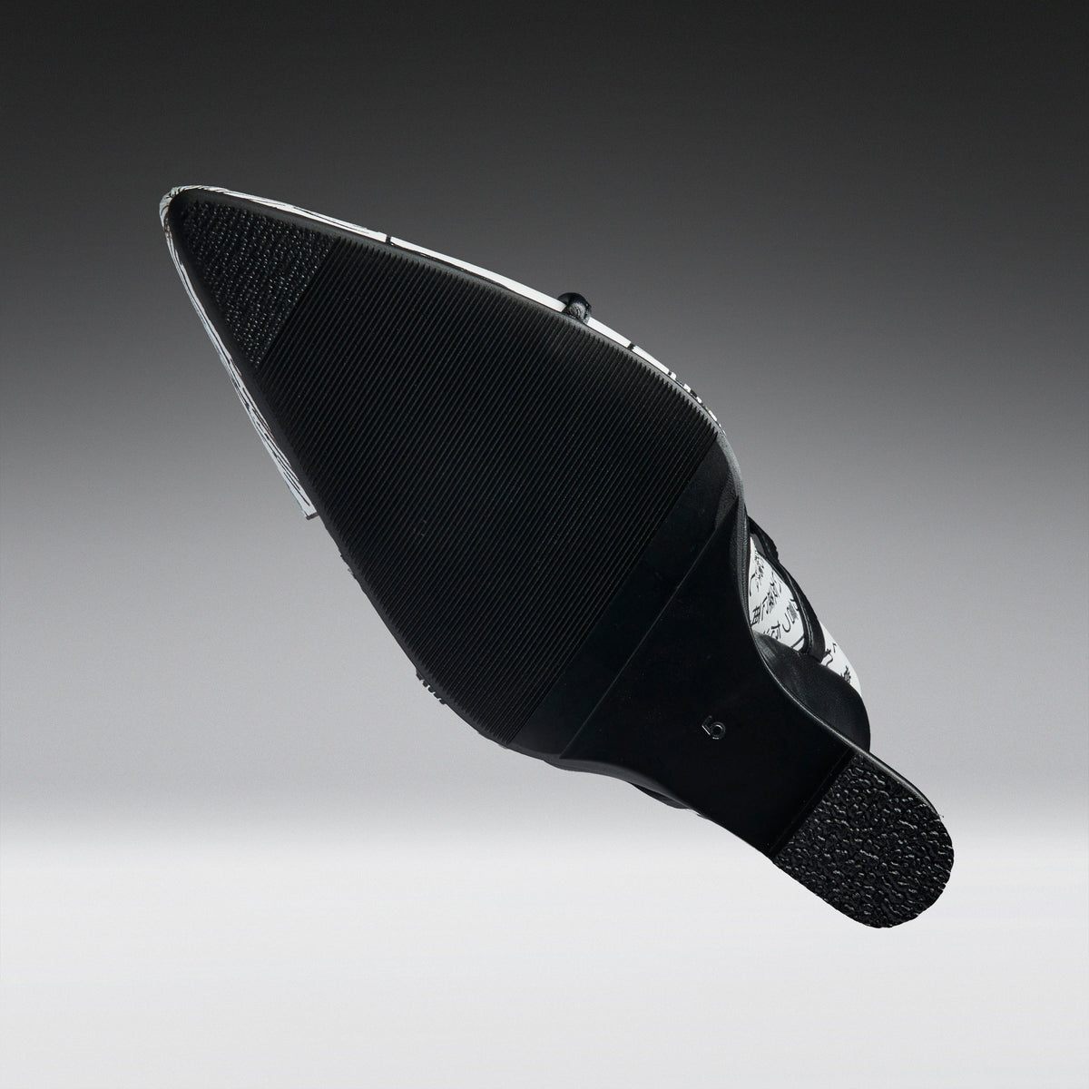 A black pointed-toe shoe shown from the sole side, featuring a textured black rubber outsole and a silver metallic side accent, set against a dark-to-light gray gradient background.