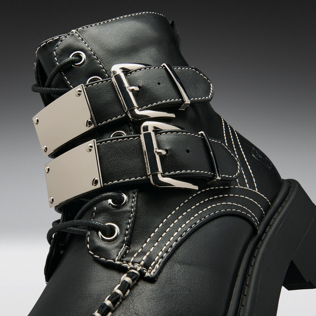 Close-up of a black leather lace-up boot with three wide decorative straps, each featuring a large silver-tone metal plate and buckle, white contrast stitching, and a chunky black sole.