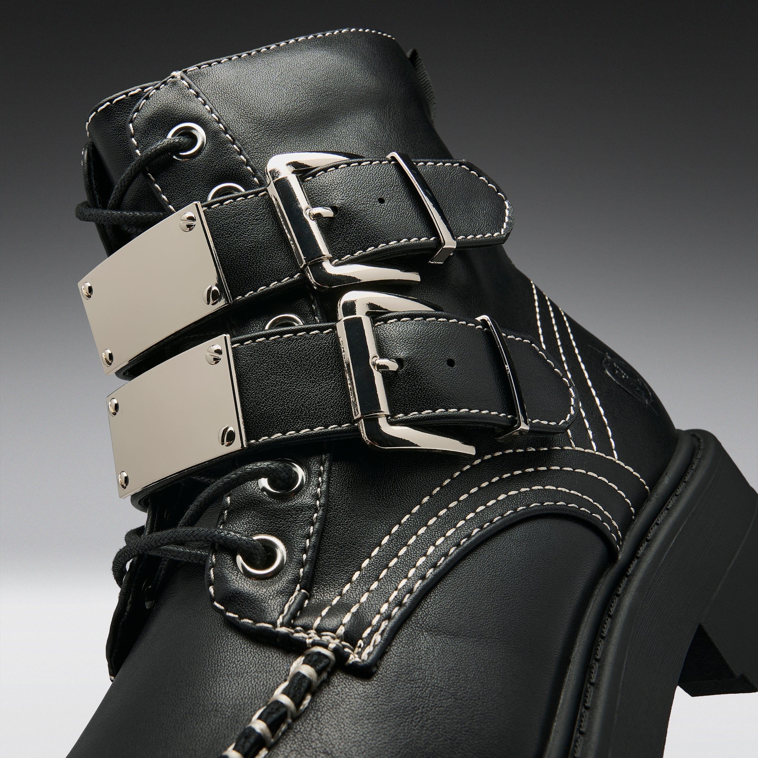 Close-up of a black leather lace-up boot with three wide decorative straps, each featuring a large silver-tone metal plate and buckle, white contrast stitching, and a chunky black sole.