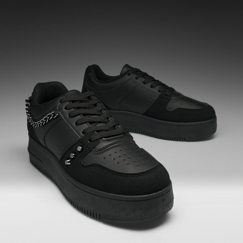 A pair of black low-top sneakers with matte and suede textures, featuring a chain detail on the side, metal studs, perforated detailing on the toe, and thick black soles, set against a dark gray gradient background.