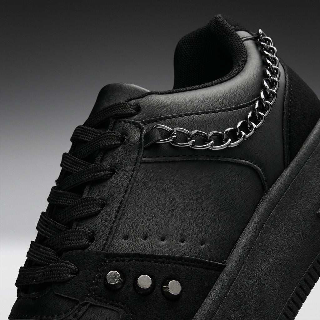 A close-up of a black low-top sneaker with black laces, a thick black sole, and a prominent silver chain draped over the side near the collar.
