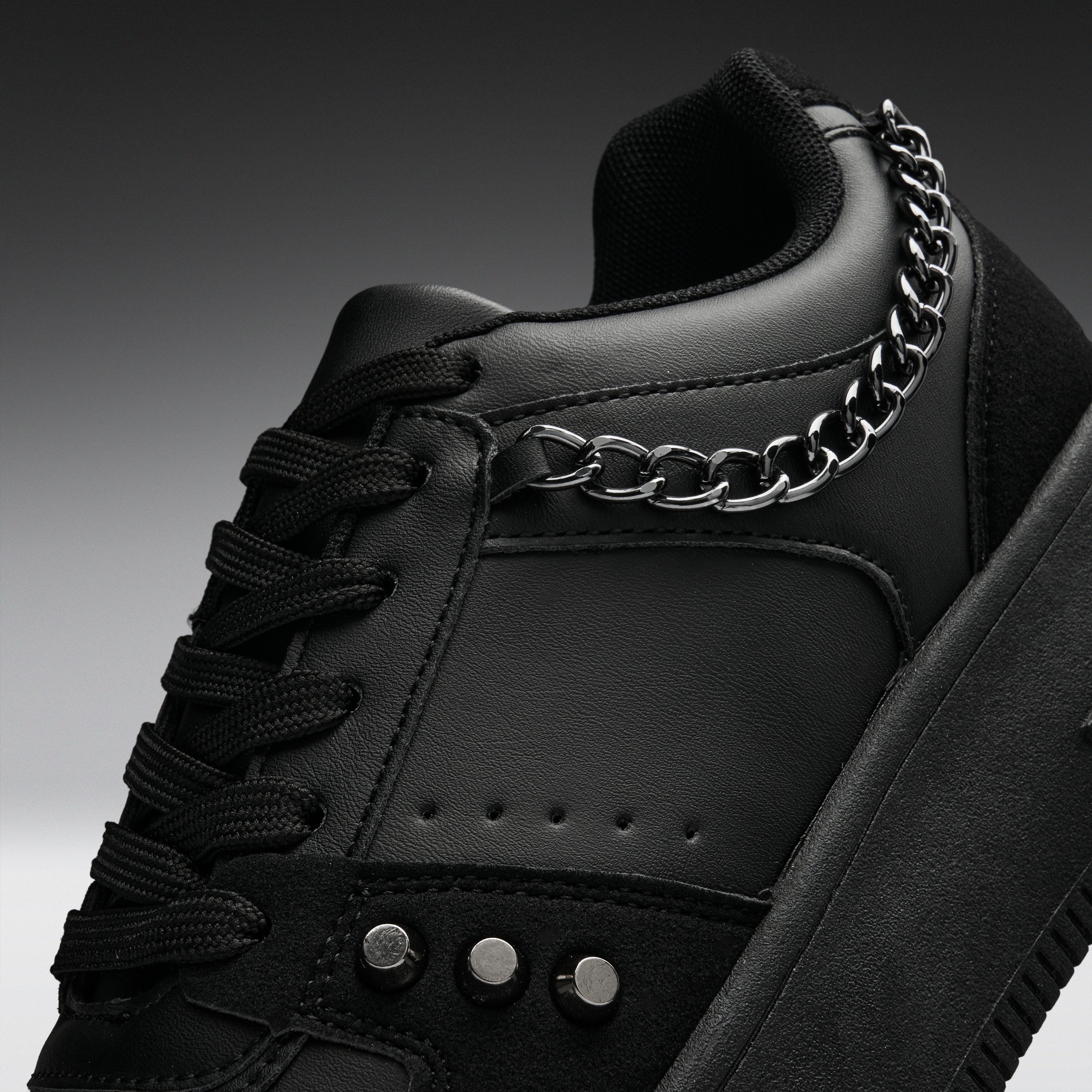 A close-up of a black low-top sneaker with black laces, a thick black sole, and a prominent silver chain draped over the side near the collar.