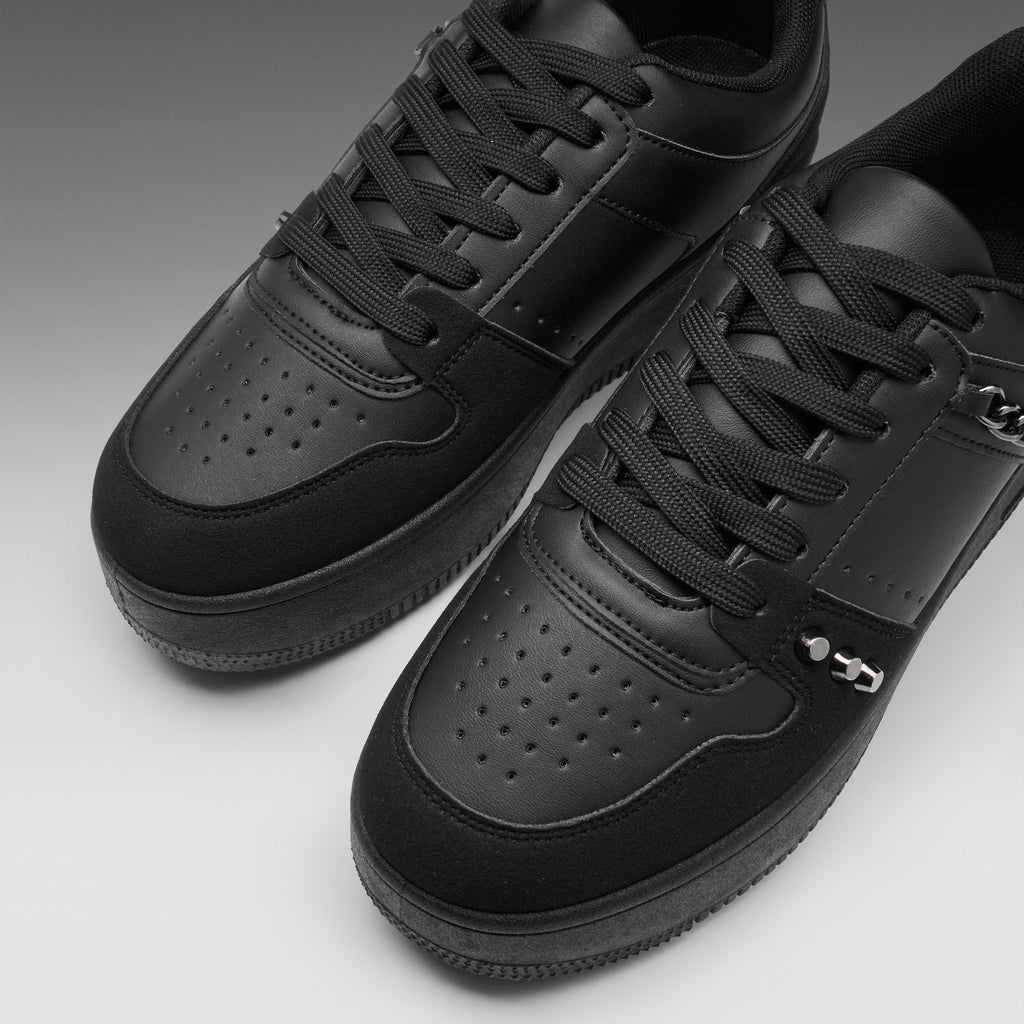 A pair of black low-top sneakers with perforated leather panels, black laces, and a small silver metal accent on the side, photographed on a clean gray studio background.
