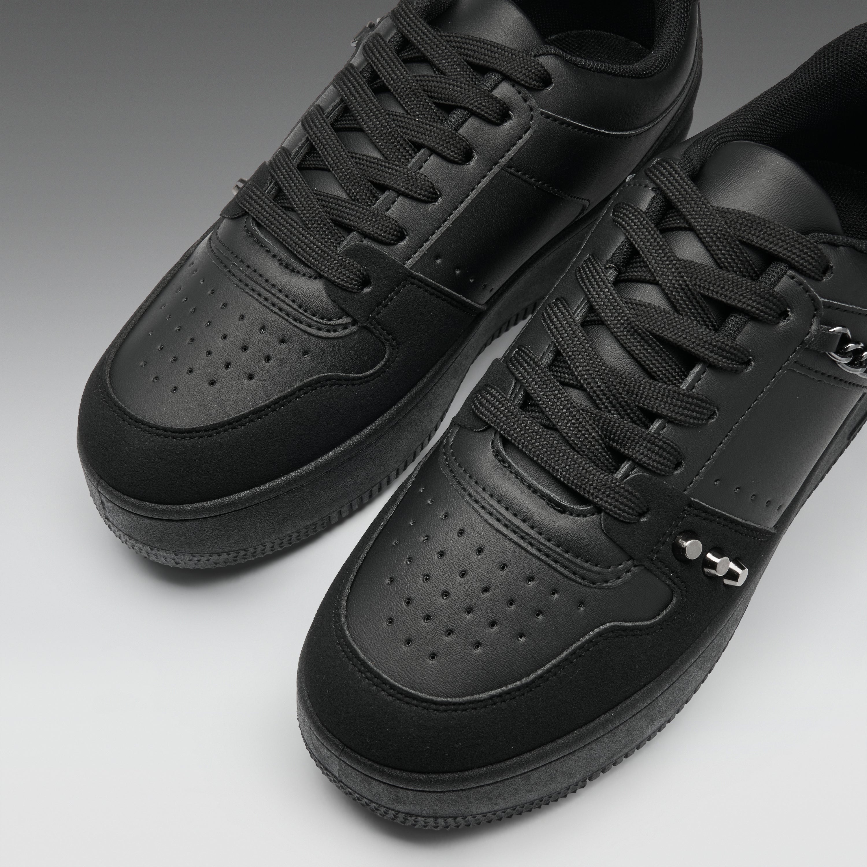 A pair of black low-top sneakers with perforated leather panels, black laces, and a small silver metal accent on the side, photographed on a clean gray studio background.