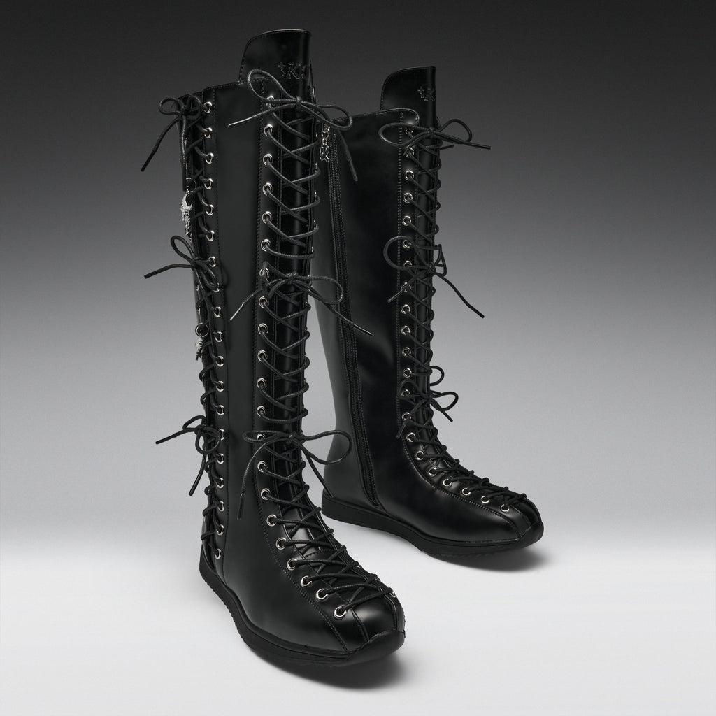 A pair of black leather knee-high lace-up boots with multiple decorative laces and silver-tone eyelets, photographed on a clean gray studio background.