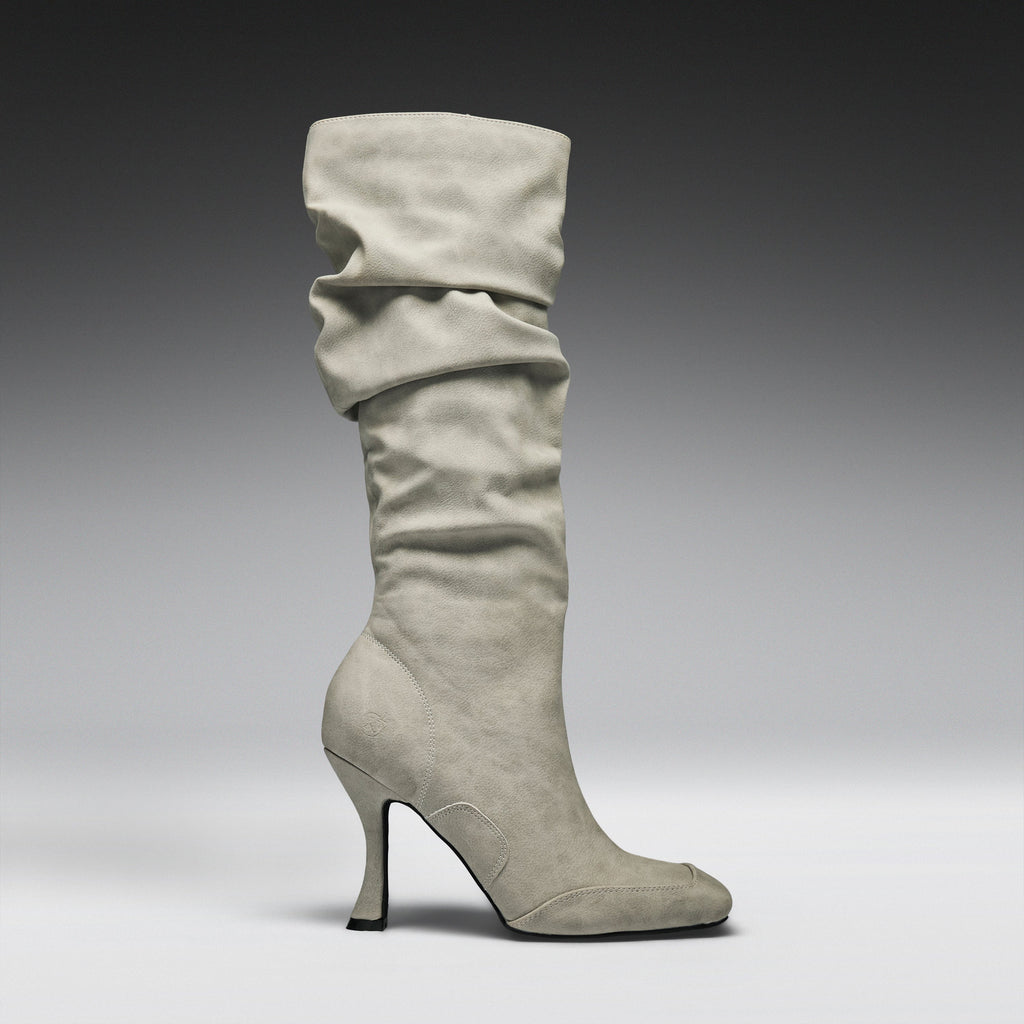 A single knee-high slouch boot in light gray suede, featuring a pointed toe and a slim stiletto heel, shown against a dark-to-light gray studio gradient background.