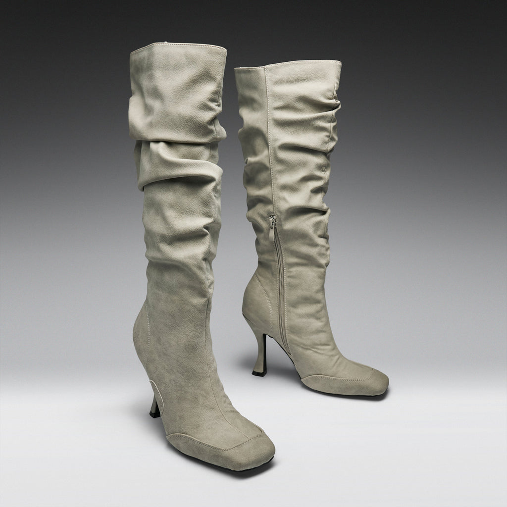 A pair of tall, knee-high heeled boots in a light taupe suede-like material, featuring a ruched shaft and a side zipper, set against a dark-to-light gray gradient studio background.