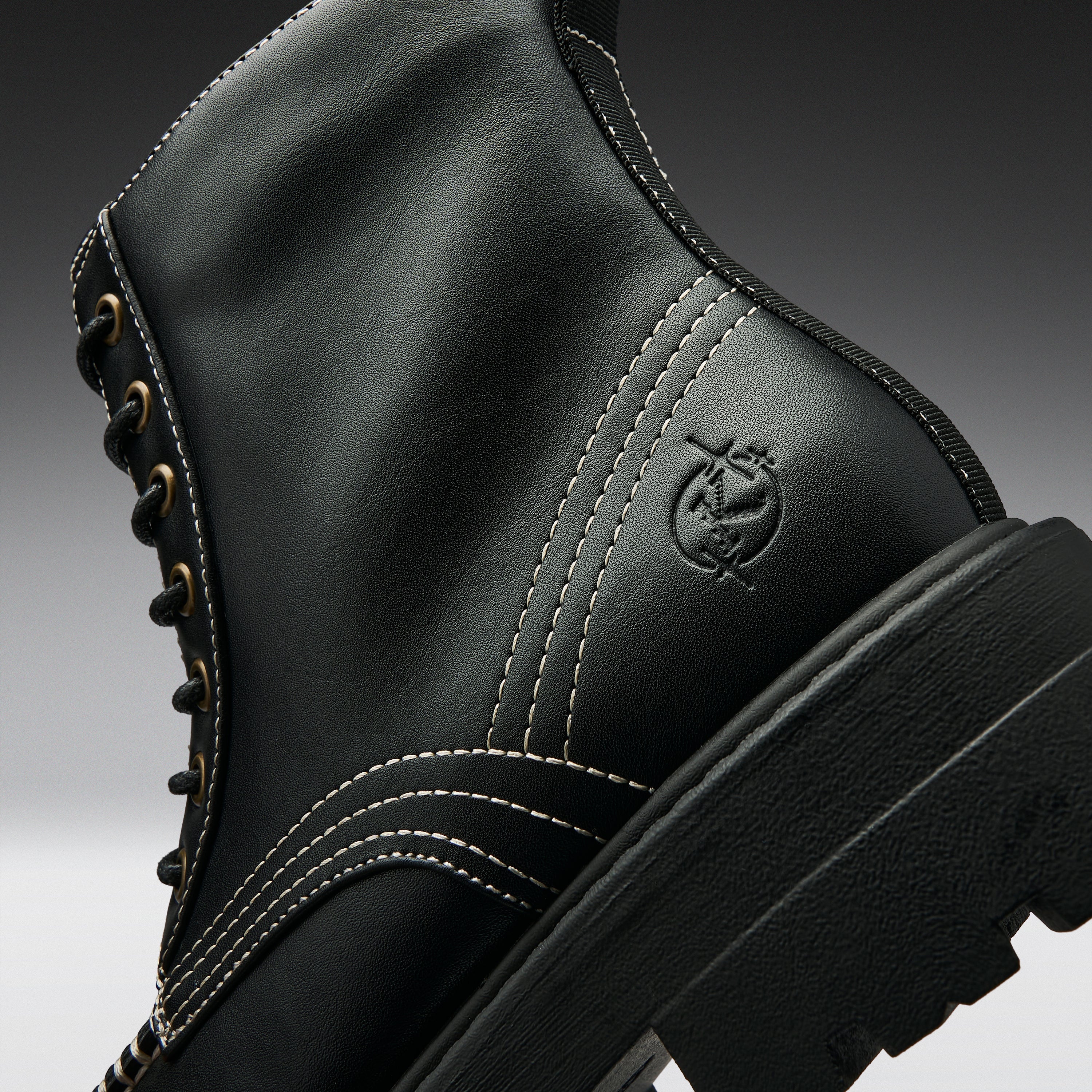 Close-up of a black leather lace-up boot with white contrast stitching, gold eyelets, and an embossed circular logo on the side, shown against a dark gray gradient background.