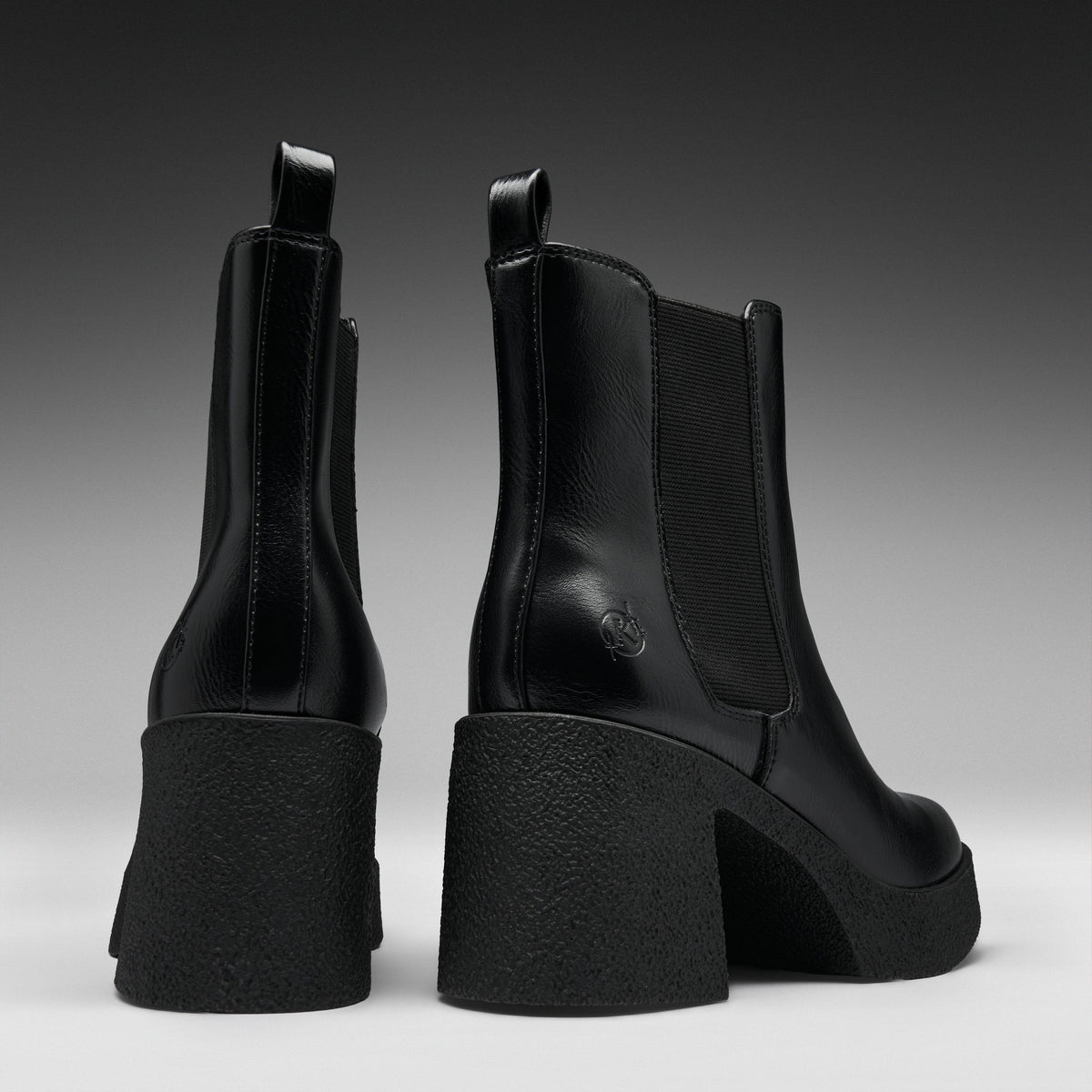 A pair of black leather Chelsea-style ankle boots with chunky block heels, shown from the back and side against a dark gray gradient studio background.