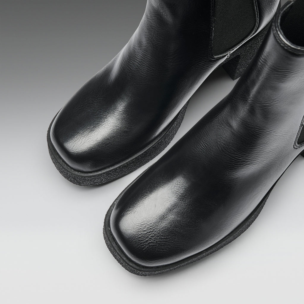 A close-up studio shot of a pair of black leather ankle boots with square toes and elastic side panels, photographed on a light gray gradient background.