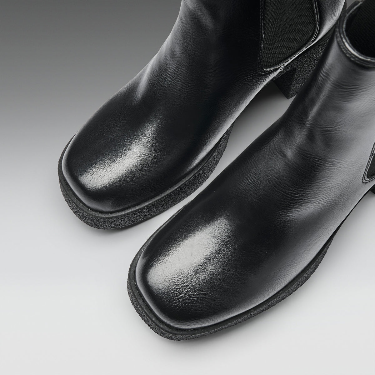 A close-up studio shot of a pair of black leather ankle boots with square toes and elastic side panels, photographed on a light gray gradient background.