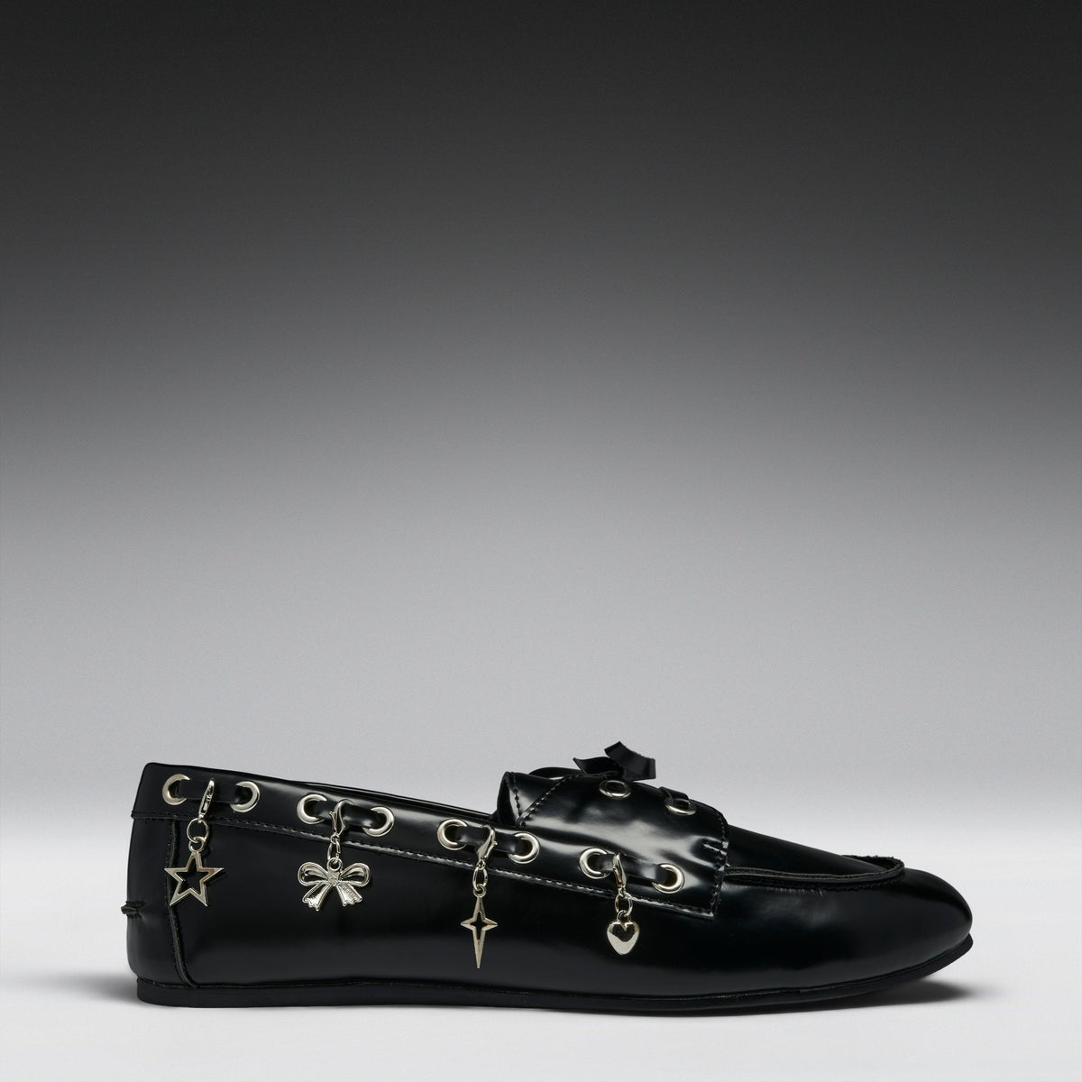 A black leather loafer flat decorated with silver-tone metal eyelets and dangling charms shaped like a star, bow, key, and heart, shown against a dark-to-light gray gradient studio background.