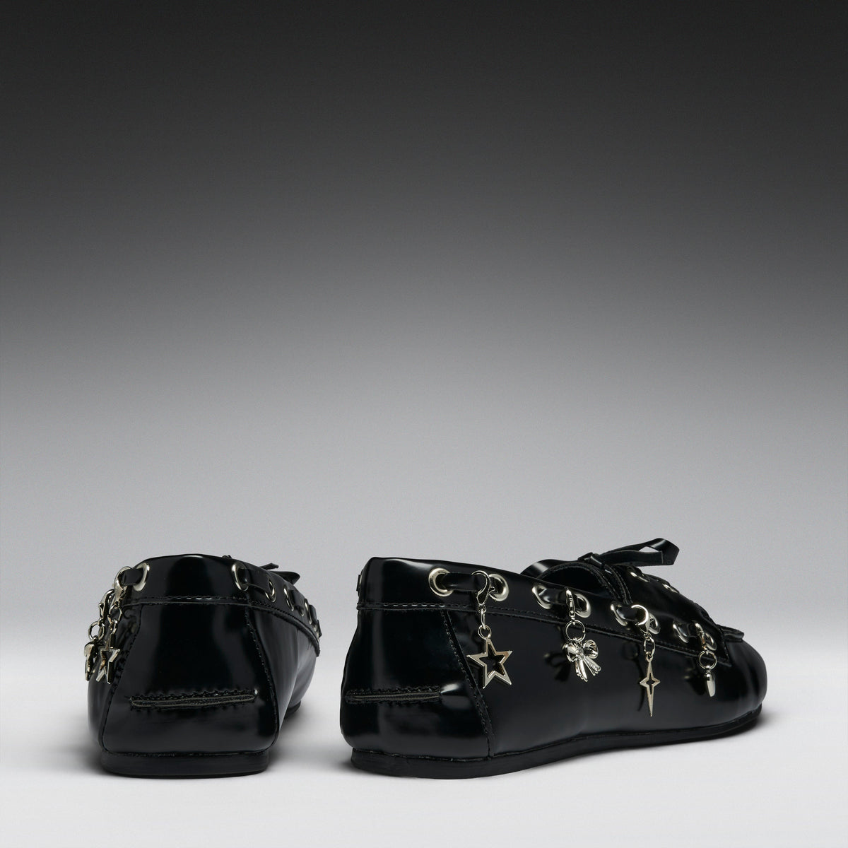 A pair of black patent leather lace-up flats decorated with silver-tone metal charms, including a star, cross, and clover, shown from the back and side against a dark gray gradient background.