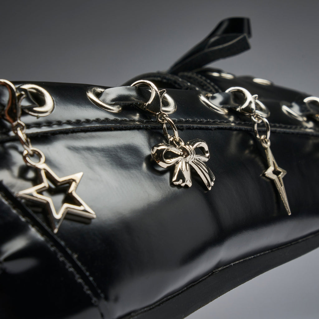 Close-up of a glossy black patent leather bag featuring silver-tone dangling charms shaped like a star, a bow, and a lightning bolt, with matching silver eyelets and studs.