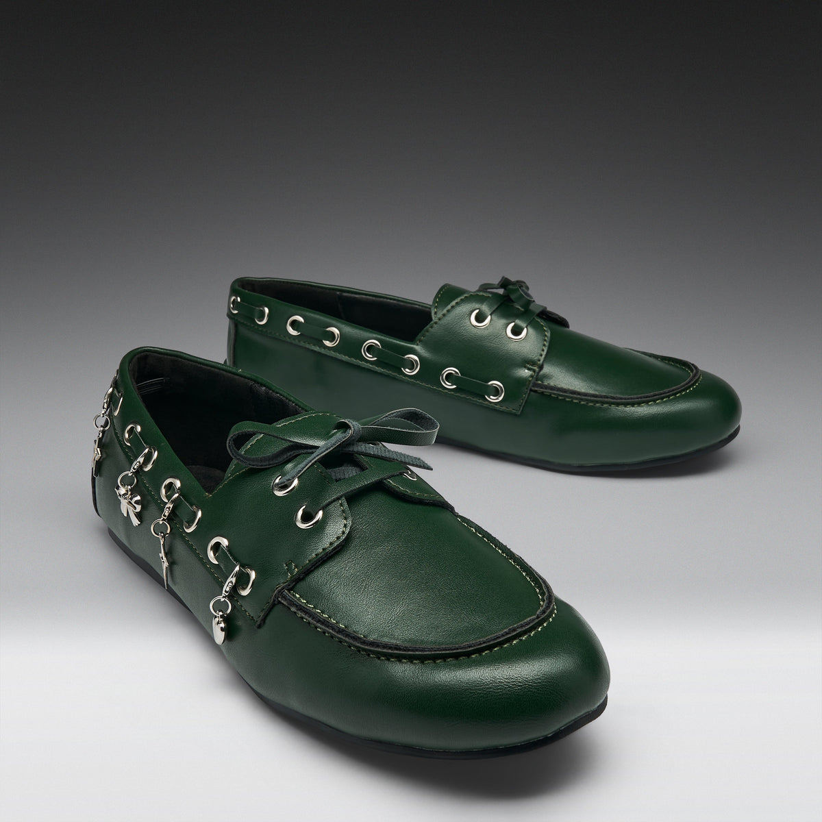 A pair of dark green leather boat-style shoes with decorative silver-tone metal eyelets, hanging charms, and matching green laces, displayed on a dark-to-light gray gradient studio background.