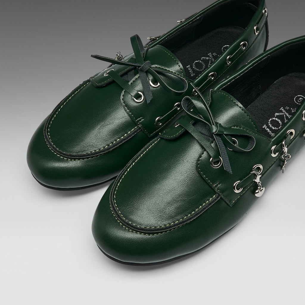 A pair of dark green leather boat-style loafers with matching green laces, silver-tone eyelets, and decorative metal charms, displayed on a neutral gray background.