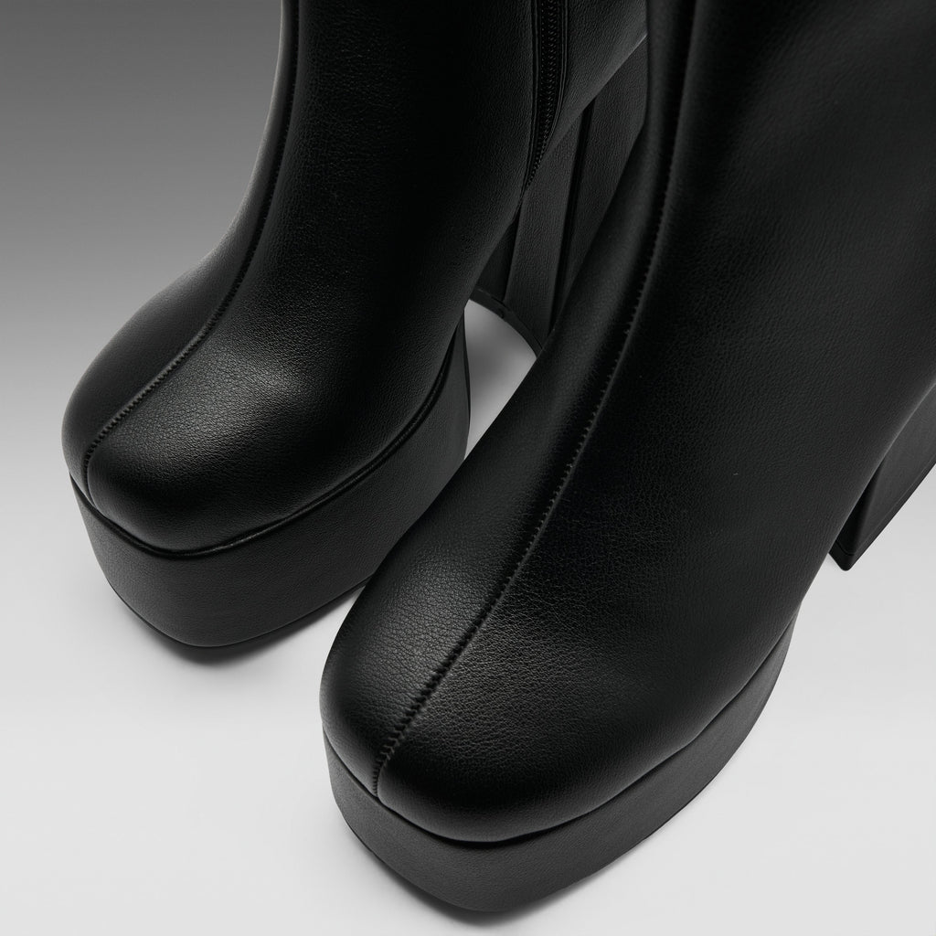 Close-up of a pair of black leather platform ankle boots with a chunky block heel, showing the toe and side profile against a neutral gray background.