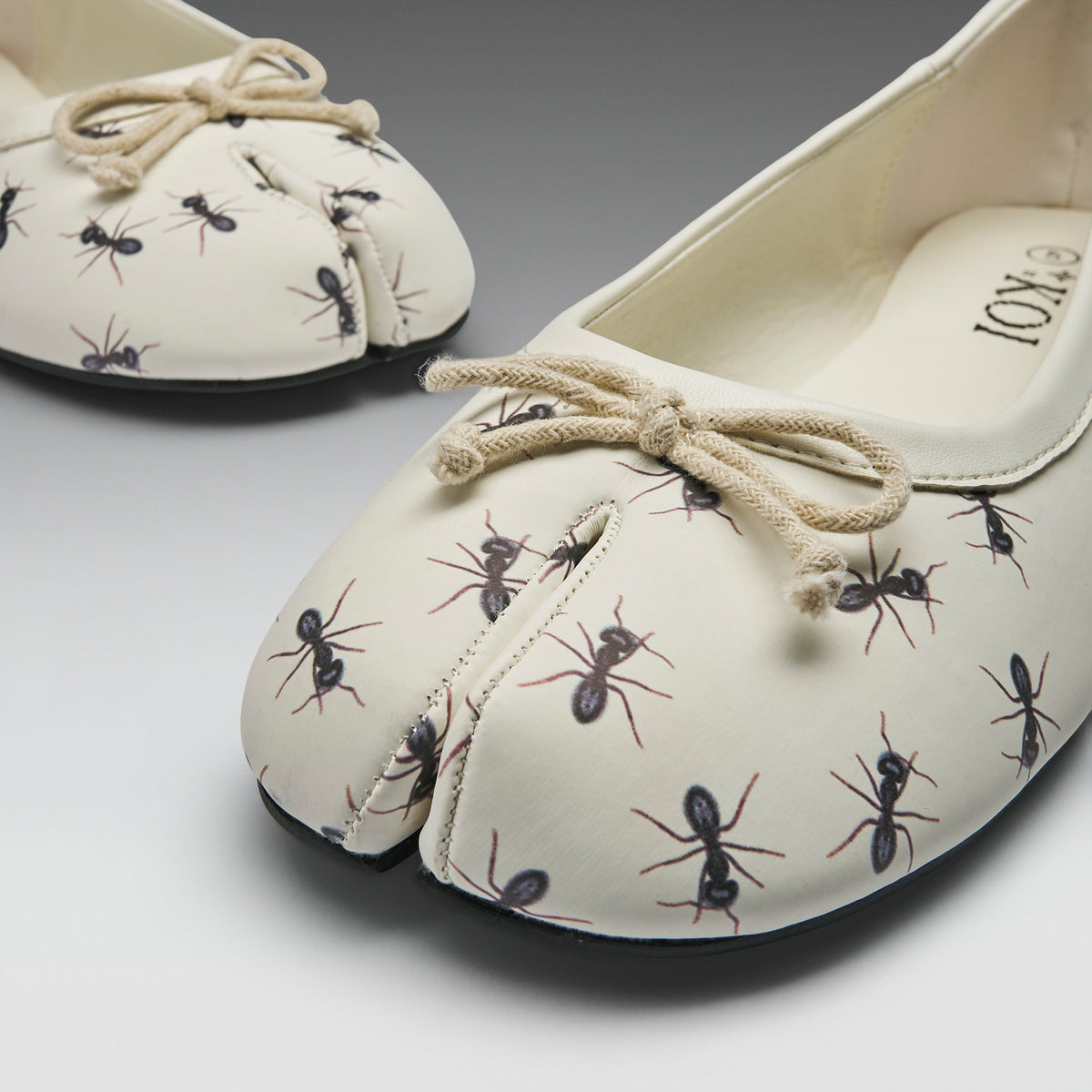 A pair of split-toe ballet flats with an off-white base, printed with black and brown ants, and finished with a beige rope bow on the top, shown on a gray gradient background.