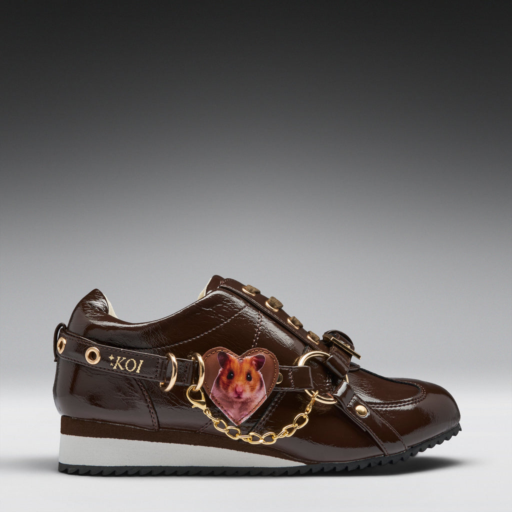 A brown patent leather sneaker featuring a heart-shaped hamster charm attached by a gold chain and strap, with 'KOI' lettering on the side.