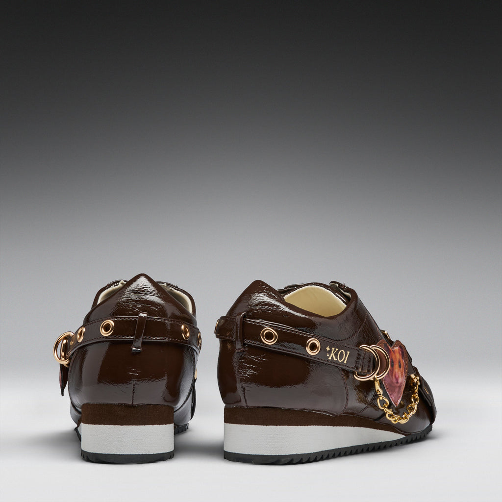 A pair of dark brown patent leather low-top sneakers shown from the back, featuring gold-tone hardware, a decorative pink and gold heart charm, and a white and black sole.