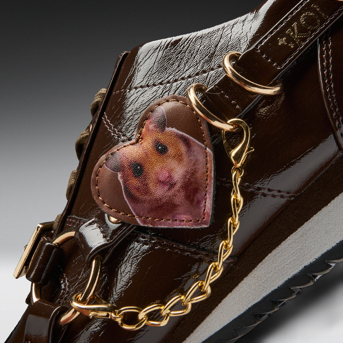 A close-up of a dark brown patent leather shoe featuring a heart-shaped leather patch with a printed hamster face, attached to gold-tone hardware and a gold chain.