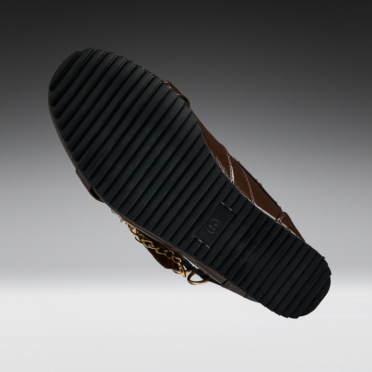 A close-up of a brown leather boot sole, showing a black ribbed rubber outsole with a small gold chain detail, set against a dark-to-light gray gradient background.