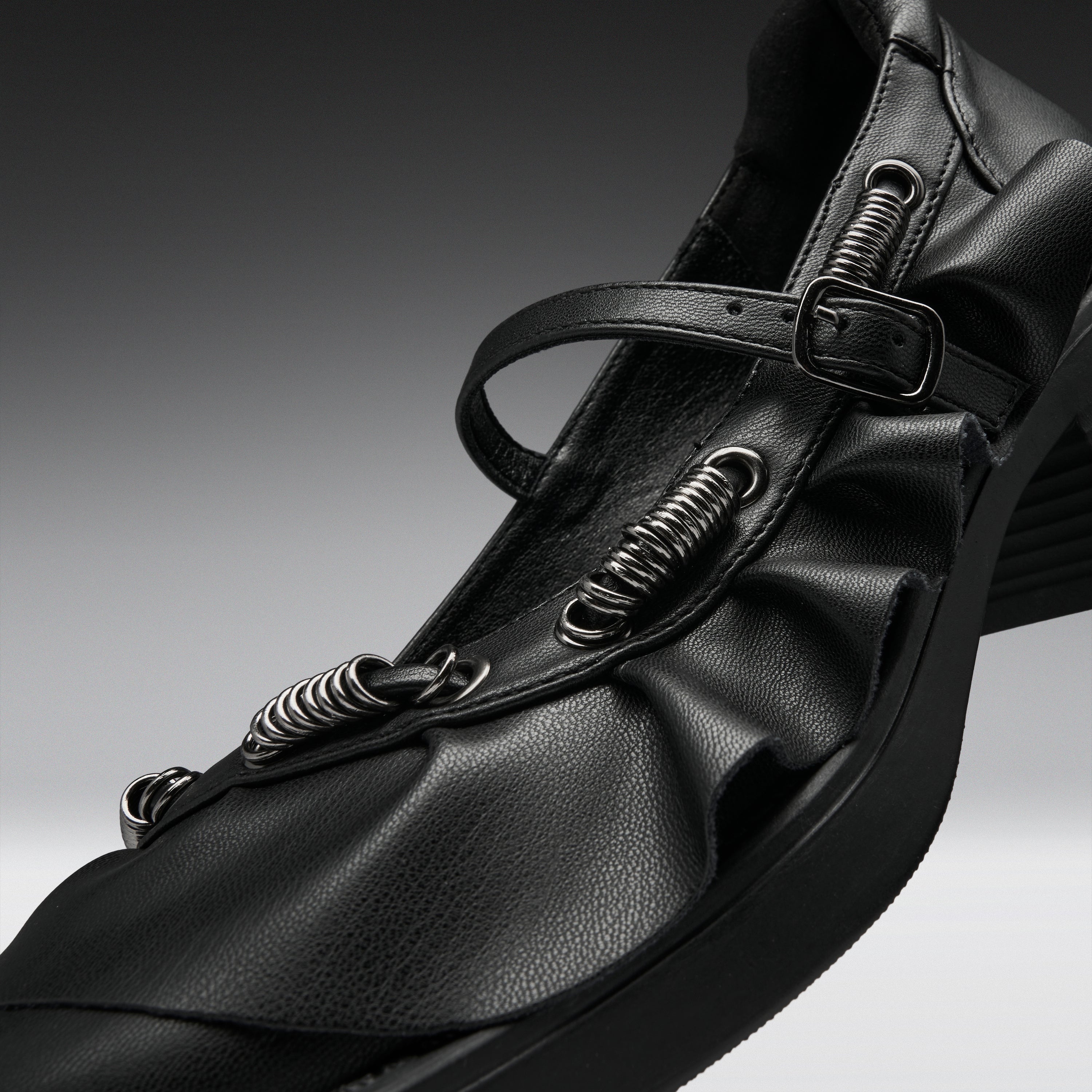 A close-up studio shot of a black leather Mary Jane-style shoe with multiple spring-loaded metal loops and a buckle strap, set against a dark gray gradient background.
