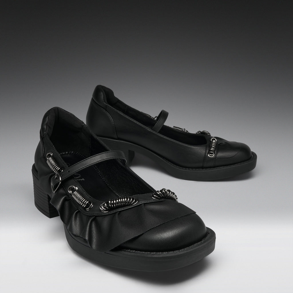 A pair of black Mary Jane-style shoes with chunky block heels, ruffled straps, and silver-tone decorative hardware, photographed on a dark-to-light gray studio background.
