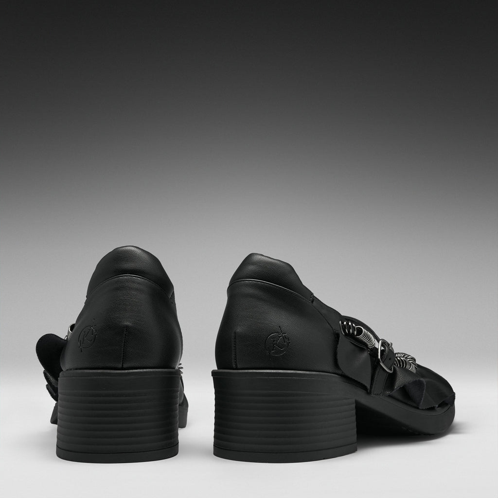 A pair of black leather high-heeled sandals shown from the back, featuring chunky stacked block heels and decorative side straps, set against a dark gray gradient background.