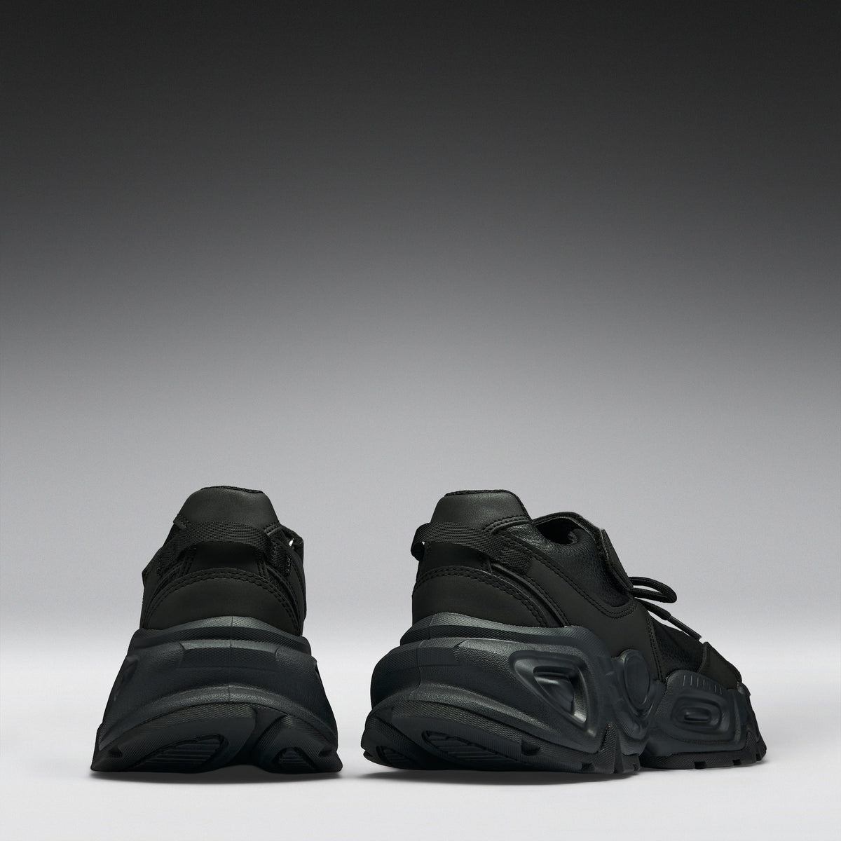 A pair of black chunky platform sneakers shown from the back, positioned side-by-side against a dark gray to light gray gradient studio background.