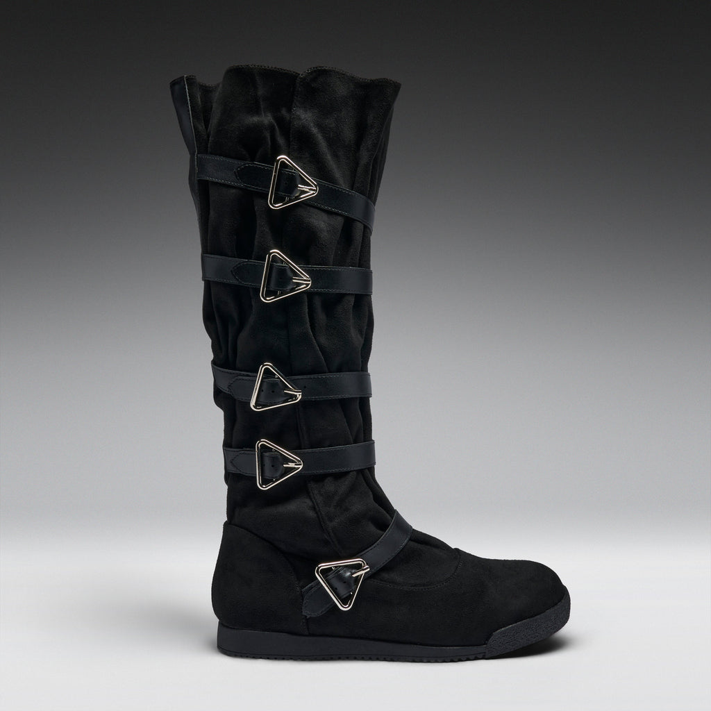 A black suede knee-high boot featuring four wrap-around straps with silver-tone triangular metal buckles, shown in profile against a dark gray gradient background.