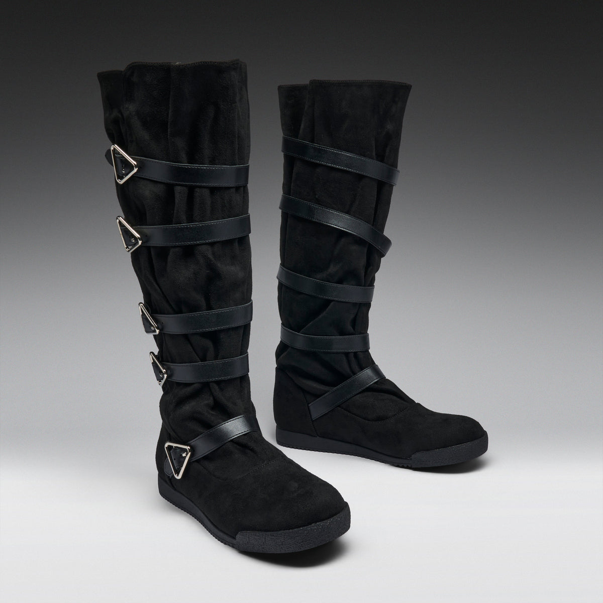 A pair of black knee-high boots with multiple wrap-around straps and silver-tone triangular buckle details, photographed on a dark-to-light gray studio background.