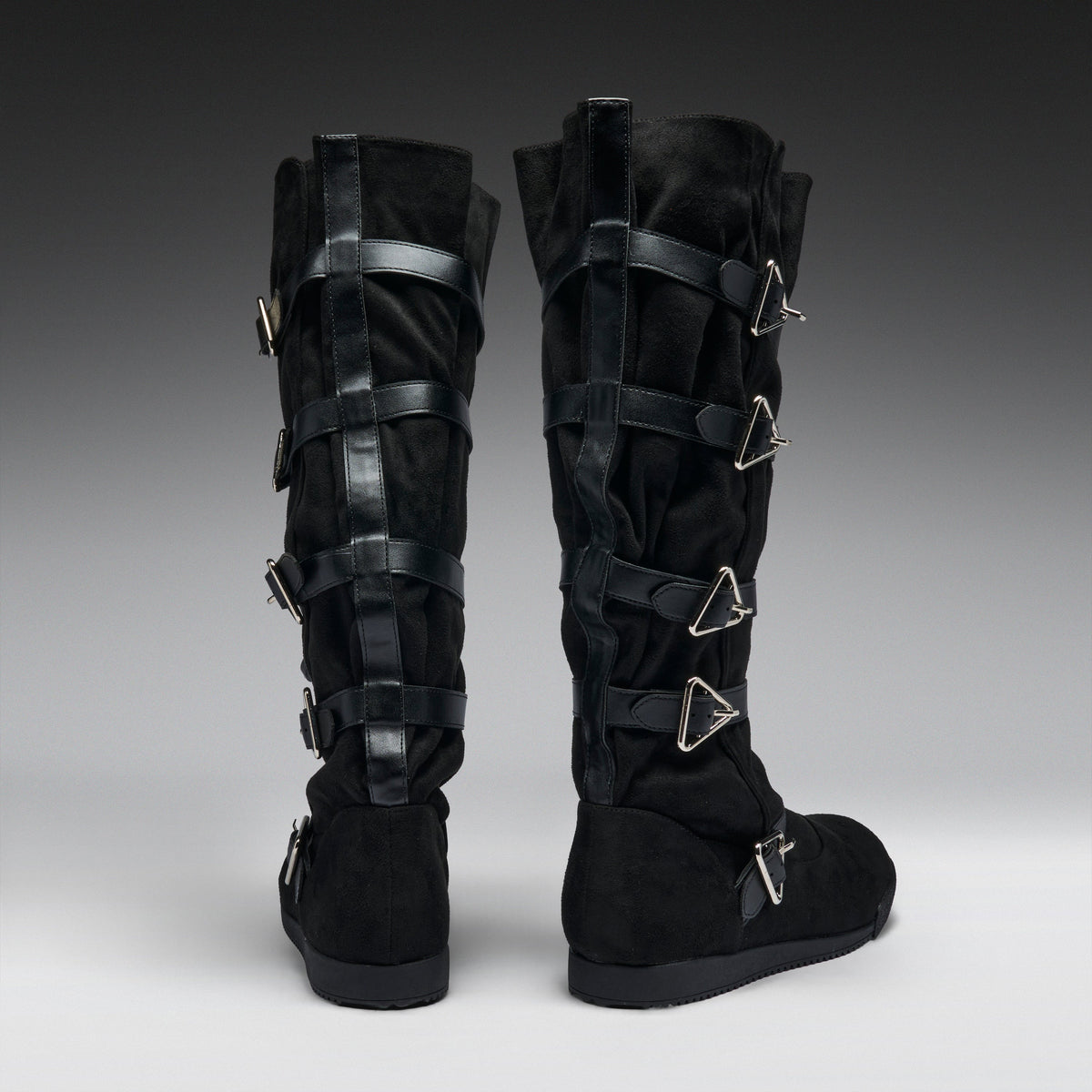 A pair of black knee-high boots with multiple black leather straps and silver-tone triangular metal buckles, shown from the back on a gray gradient studio background.