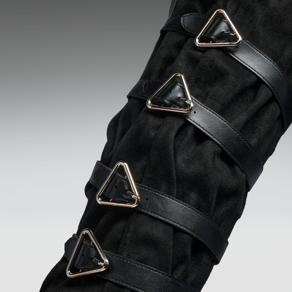 A close-up of a black textured boot or leg cover with multiple black straps and shiny silver triangular metal buckles, shown against a neutral gray gradient background.