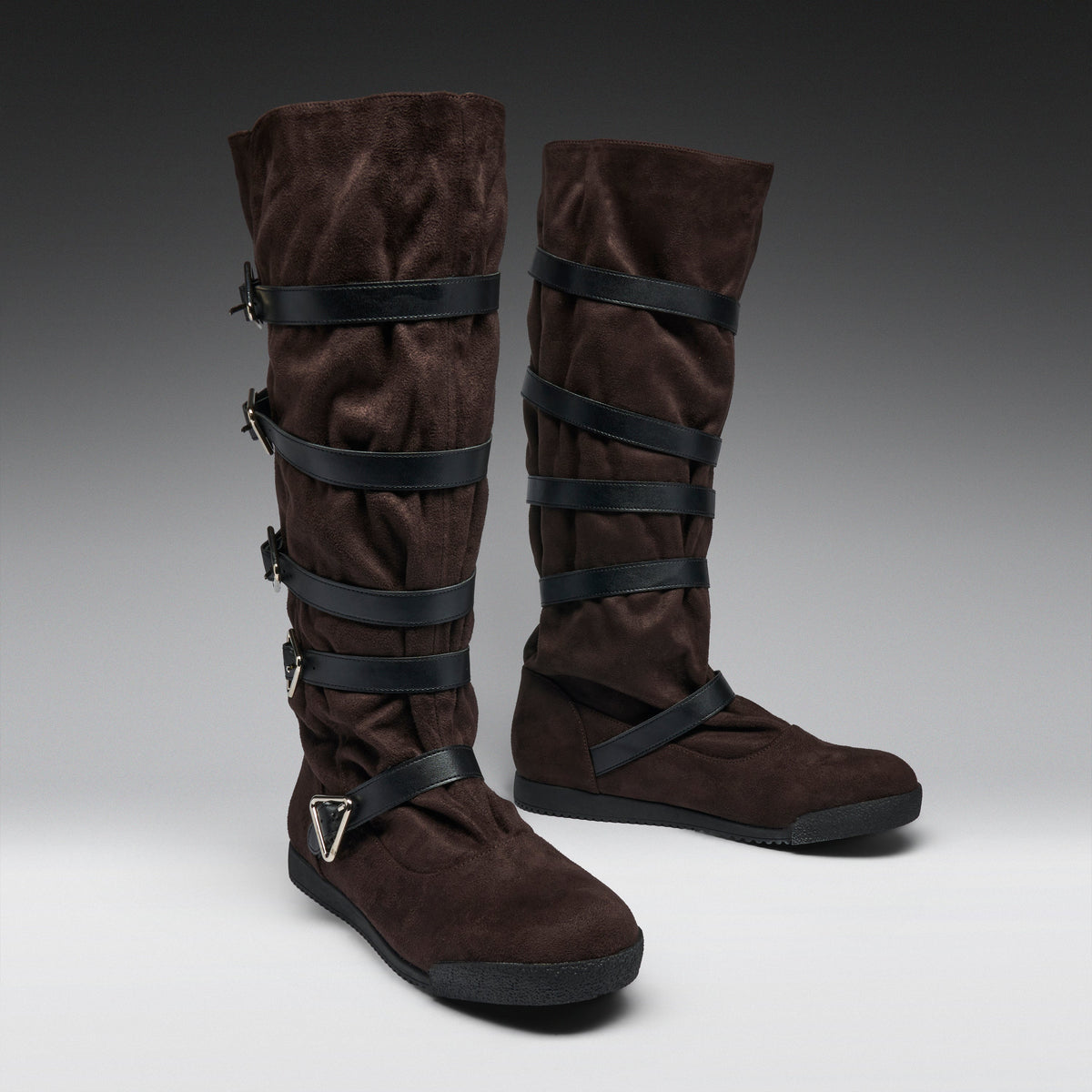 A pair of tall brown suede boots with multiple black wrap-around straps and buckles, displayed on a neutral gray studio background.
