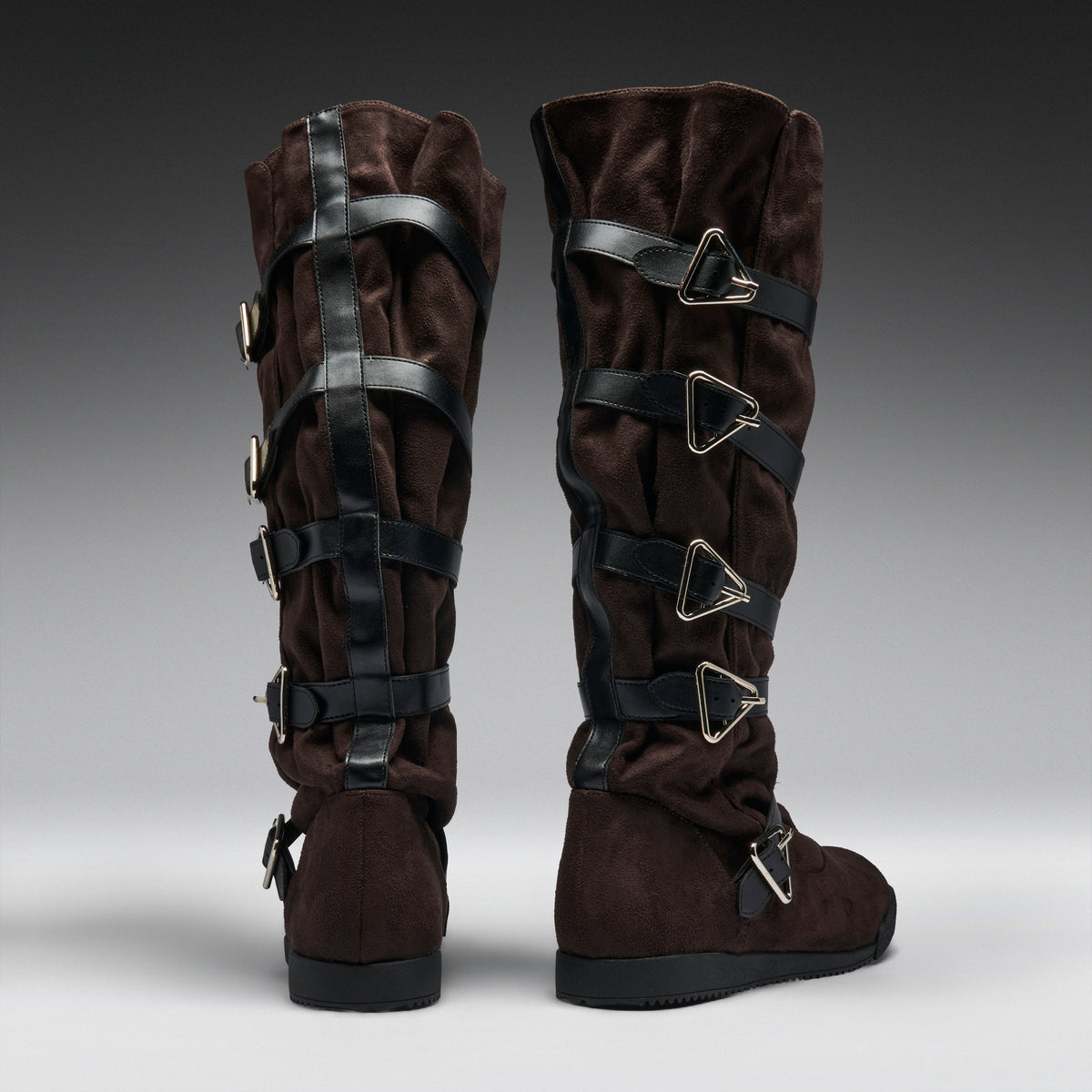 A pair of dark brown suede knee-high boots with multiple black faux-leather straps and silver-tone triangular buckle details, shown from the back on a gray gradient studio background.