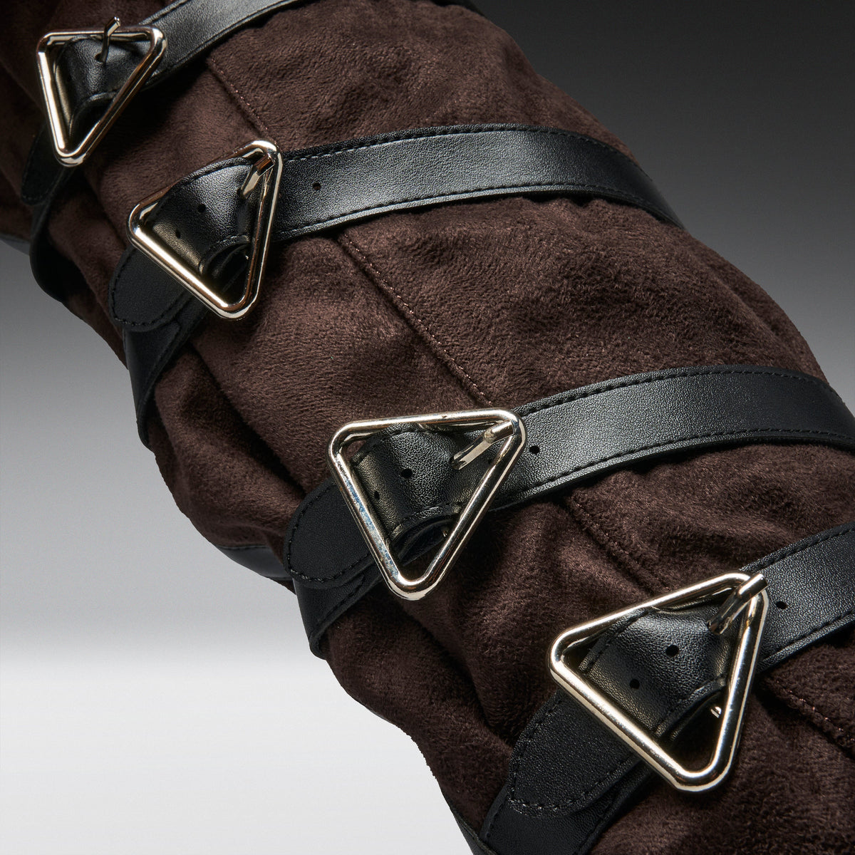 Close-up of a dark brown suede or faux-suede garment with multiple black leather straps and large triangular silver metal buckles.